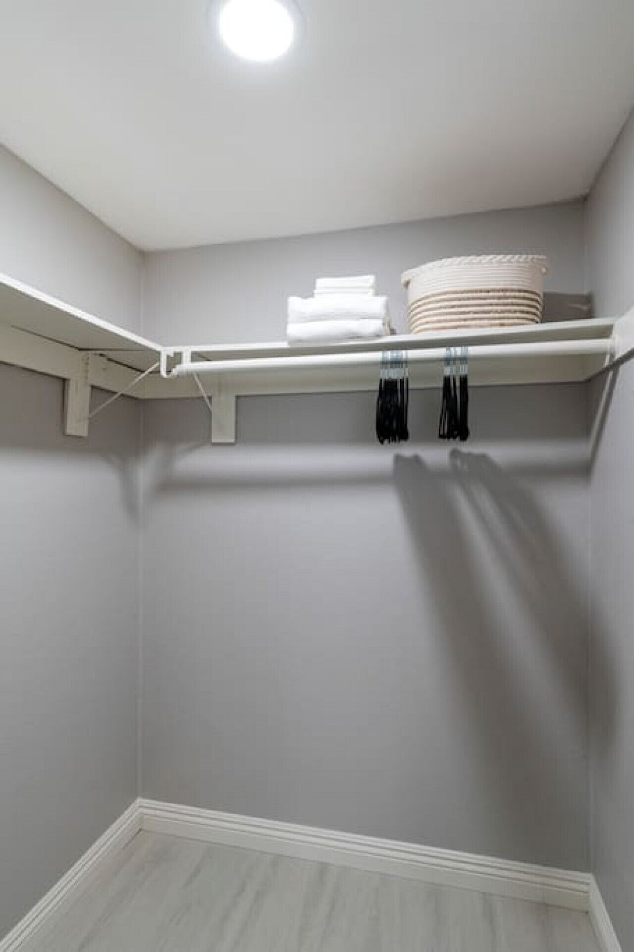 Un vestidor con pared gris, techo blanco y suelo de madera. Hay dos perchas negras y una pila de toallas dobladas sobre un estante blanco.