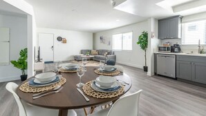 Un soggiorno moderno e luminoso con zona pranzo apparecchiata per un pasto, un divano grigio e una cucina con mobili ed elettrodomestici grigi.