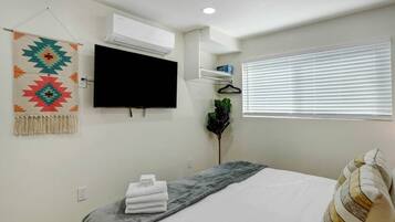 Un dormitorio con una cama, televisión, aire acondicionado, ventana con persianas y un cuadro en la pared.