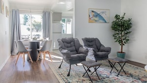 A modern living room with a dining area, grey upholstered chairs, a potted plant, and a colorful rug.