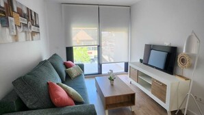 Ein modernes Wohnzimmer mit grauem Sofa, einem Holztisch, einem Flachbildfernseher auf einem weißen TV-Schrank und einem großen Fenster mit Jalousien.