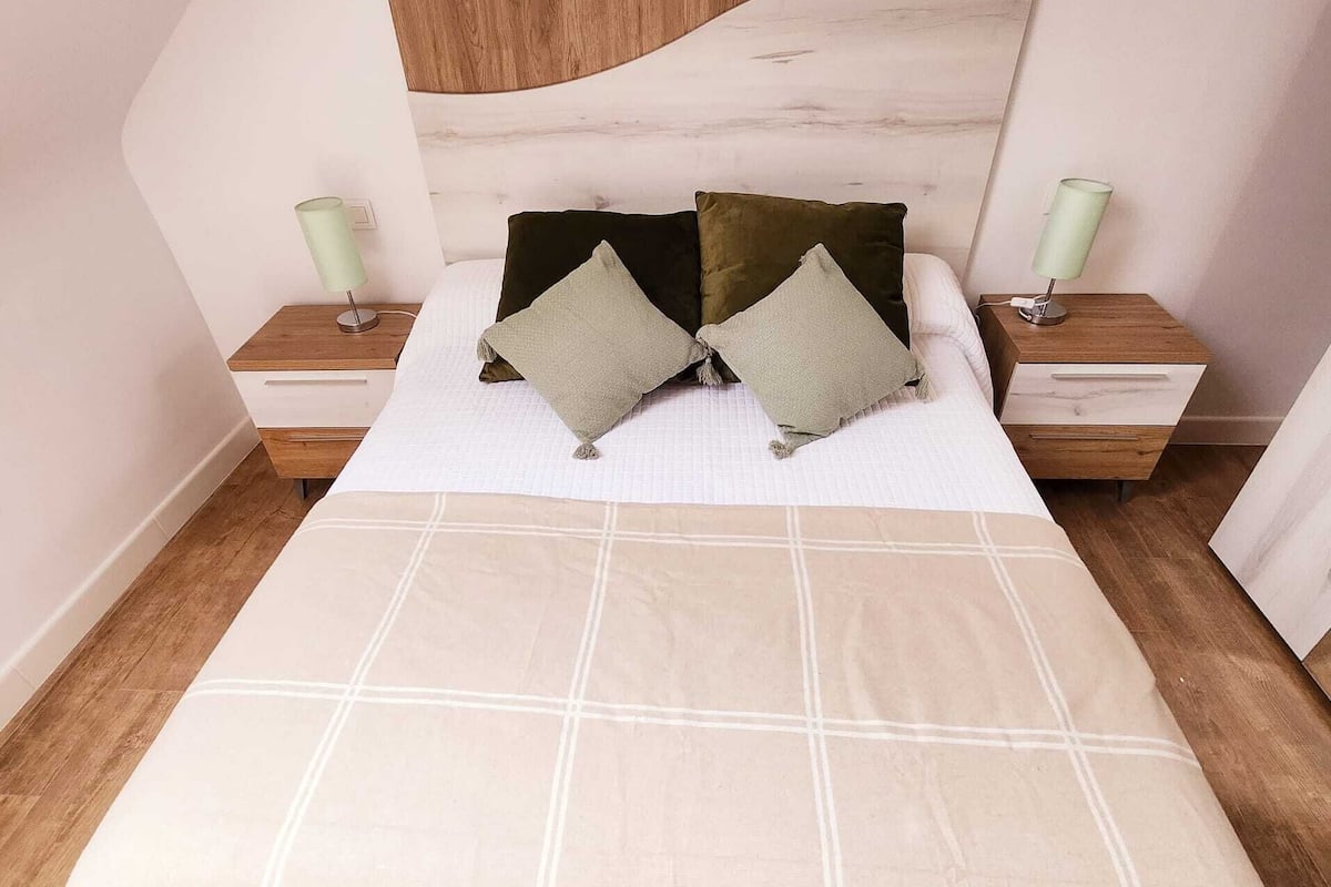 A neatly made bed with a beige bedspread, two pillows, and a wooden headboard. There are two bedside tables with lamps on either side of the bed.