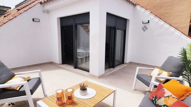 Eine Dachterrasse mit einem weißen Tisch, zwei Stühlen, einer Couch mit bunten Kissen und einem Topfpflanzen.