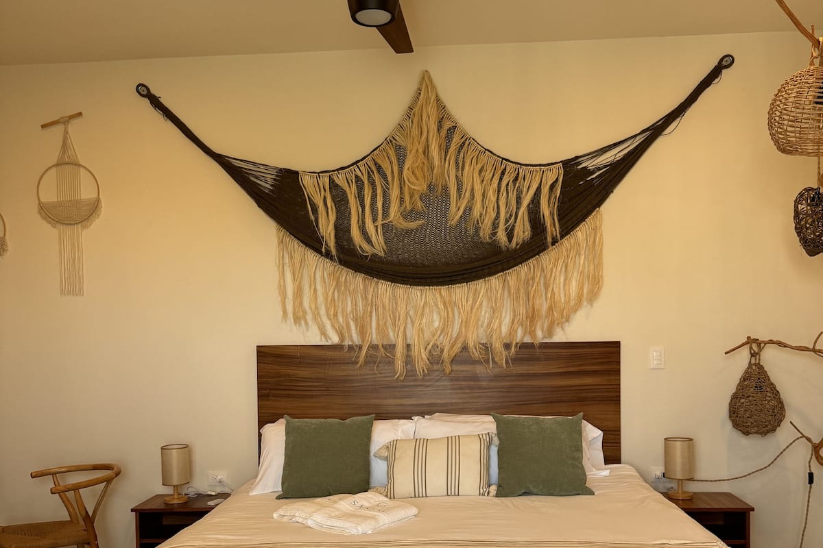 Een net opgemaakt bed met een houten hoofdbord, een plafondventilator en een hangmat bevestigd aan het plafond.