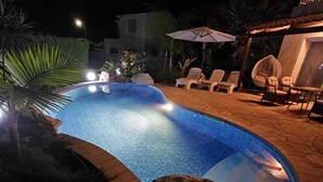 Una piscina iluminada de noche con agua cristalina, rodeada de sillones de descanso y sombrillas, con rocas grandes al frente.