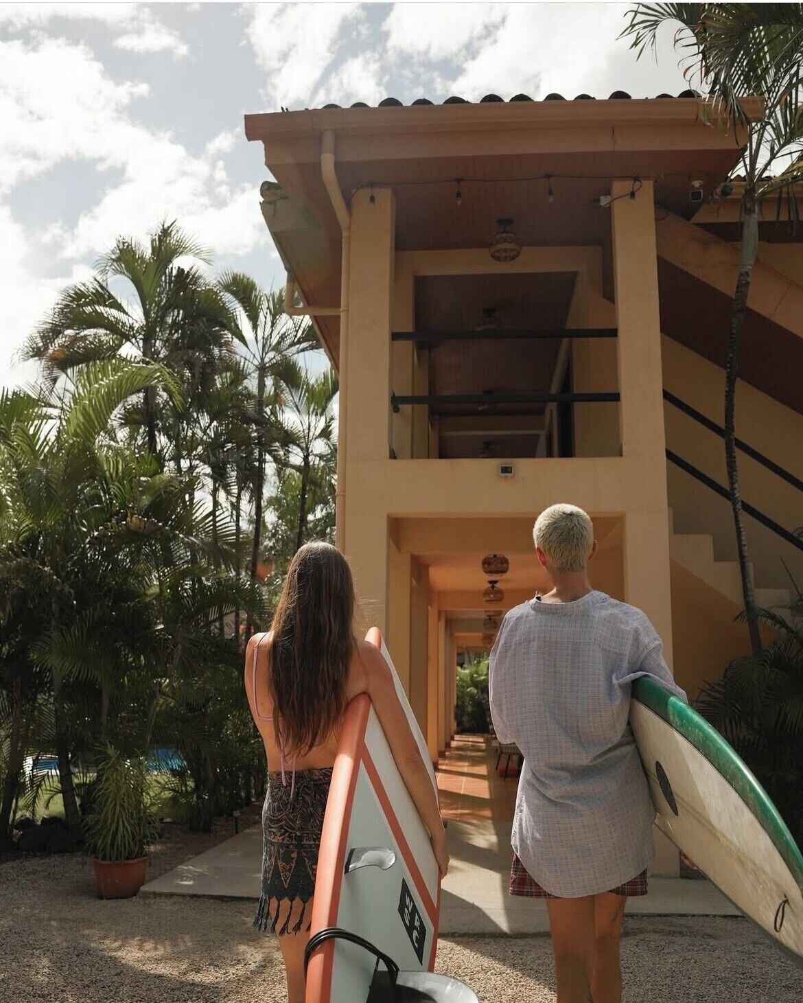 Two people carrying surfboards, walking towards a building with a tiled roof and a staircase.
