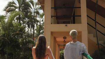 Two people carrying surfboards, walking towards a building with a tiled roof and a staircase.