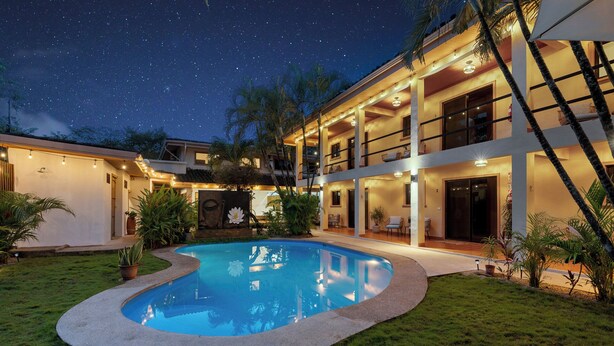 Un edificio de dos pisos con piscina, iluminación exterior y un cielo nocturno estrellado.