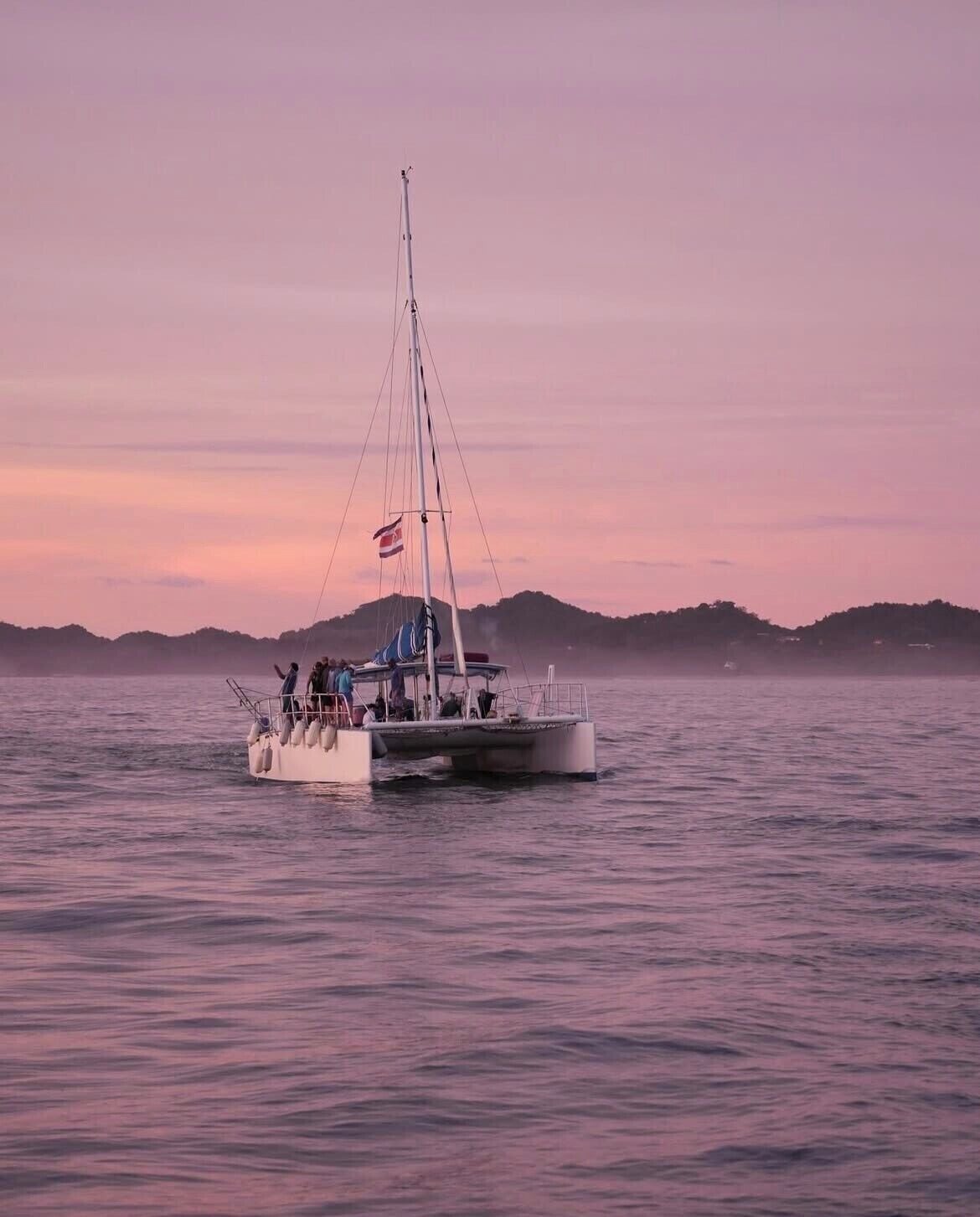 A sailboat with people on board, anchored in calm waters with a hilly landscape in the background.