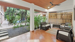 Un espacio interior amplio con techo de madera, un área de descanso con mobiliario de mimbre y vistas a un jardín tropical.