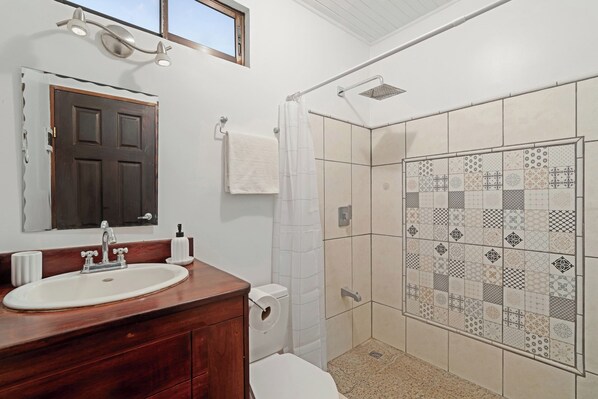Un baño con tocador de madera, un lavabo blanco, un espejo, toallero y una ducha con azulejos estampados, además de una ventana sobre el espejo.