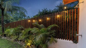 Vista nocturna de un jardín con valla de madera, palmeras y luces colgantes.