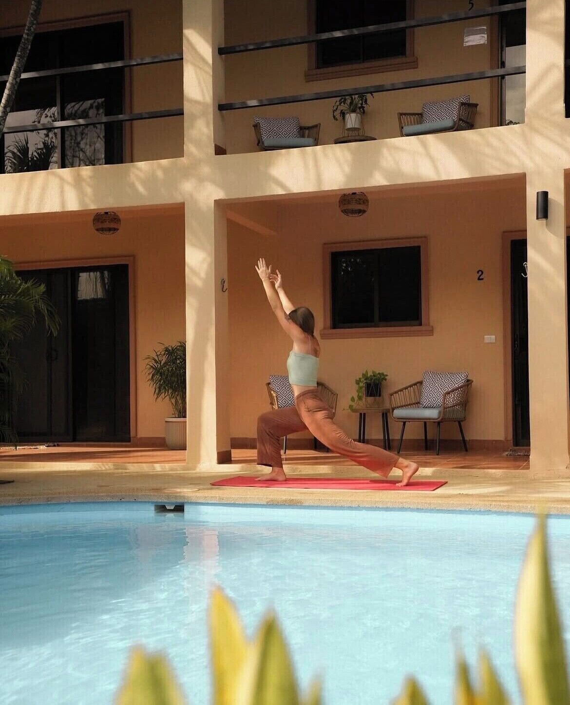 Una persona realizando una postura de yoga sobre una colchoneta junto a una piscina, en un edificio con balcones y plantas en macetas.