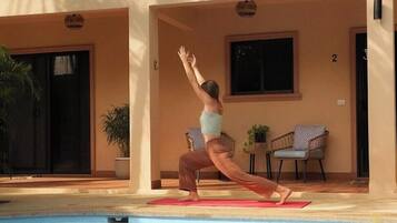 Una persona realizando una postura de yoga sobre una colchoneta junto a una piscina, en un edificio con balcones y plantas en macetas.