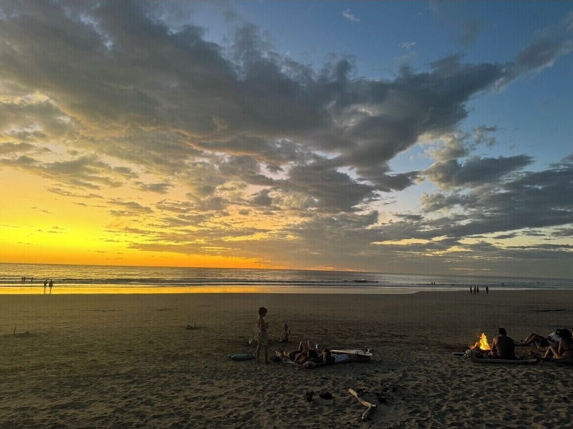 Una playa al atardecer con gente relajándose y una fogata en la arena.