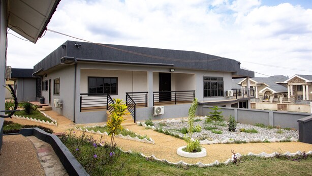 A modern two-story house with a flat roof, a covered patio, and a landscaped garden with gravel pathways and various plants. - LC Lodge & Event Centre (Kumasi)