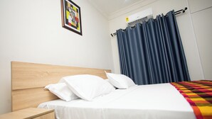 A neatly made bed with white linens and a colorful blanket, a wooden nightstand with a drawer, a wall-mounted air conditioner, and a framed picture on the wall. - LC Lodge & Event Centre (Kumasi)