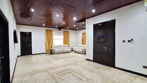 A spacious living room with a beige sofa set, a large window with yellow curtains, and a wooden ceiling with recessed lighting. - LC Lodge & Event Centre (Kumasi)
