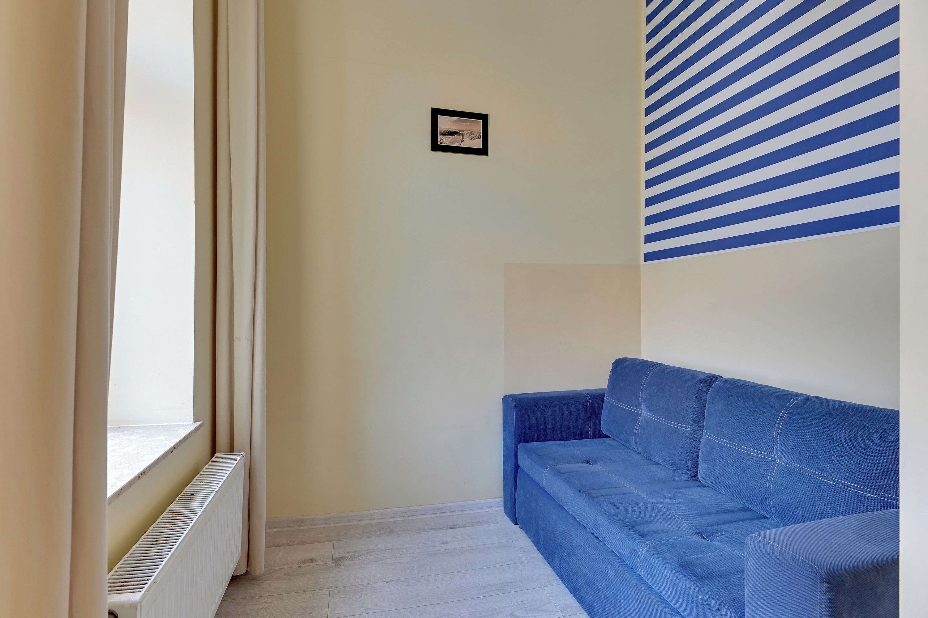 A room with a blue sofa, a radiator, a window with curtains, and a wall with a striped pattern.