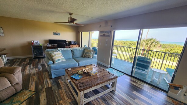 A living room with a blue sofa, wooden coffee table, and a view of the ocean through large sliding glass doors. - Direct Beachfront Sea Oats Condo in Cocoa Beach (Cocoa Beach)