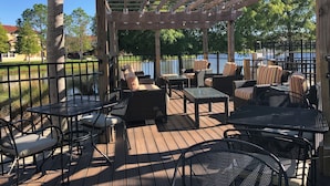 Une terrasse extérieure aménagée avec des meubles en osier, des tables et des chaises en métal, donnant sur une étendue d’eau.