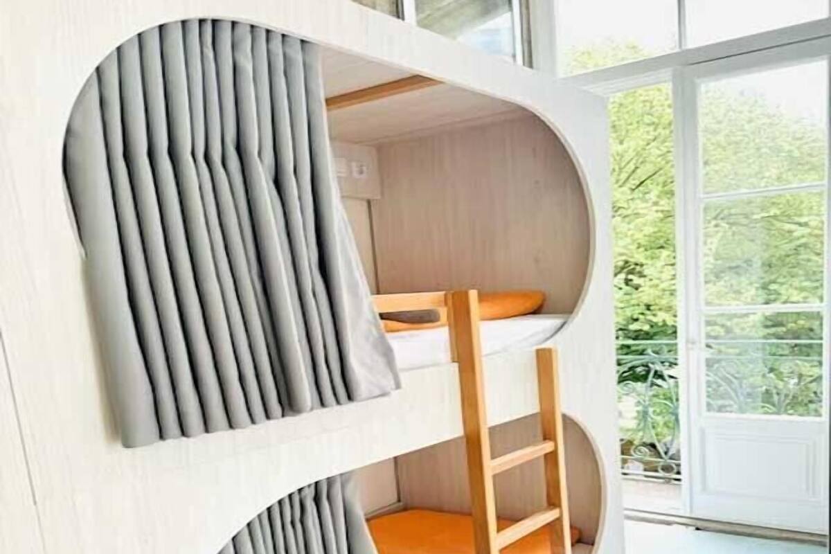 A bunk bed with a ladder, grey curtains, and a view of greenery outside.