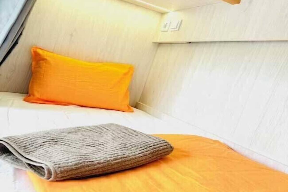 A small, compact room with a single bed, a bright orange pillow, and a folded blanket.