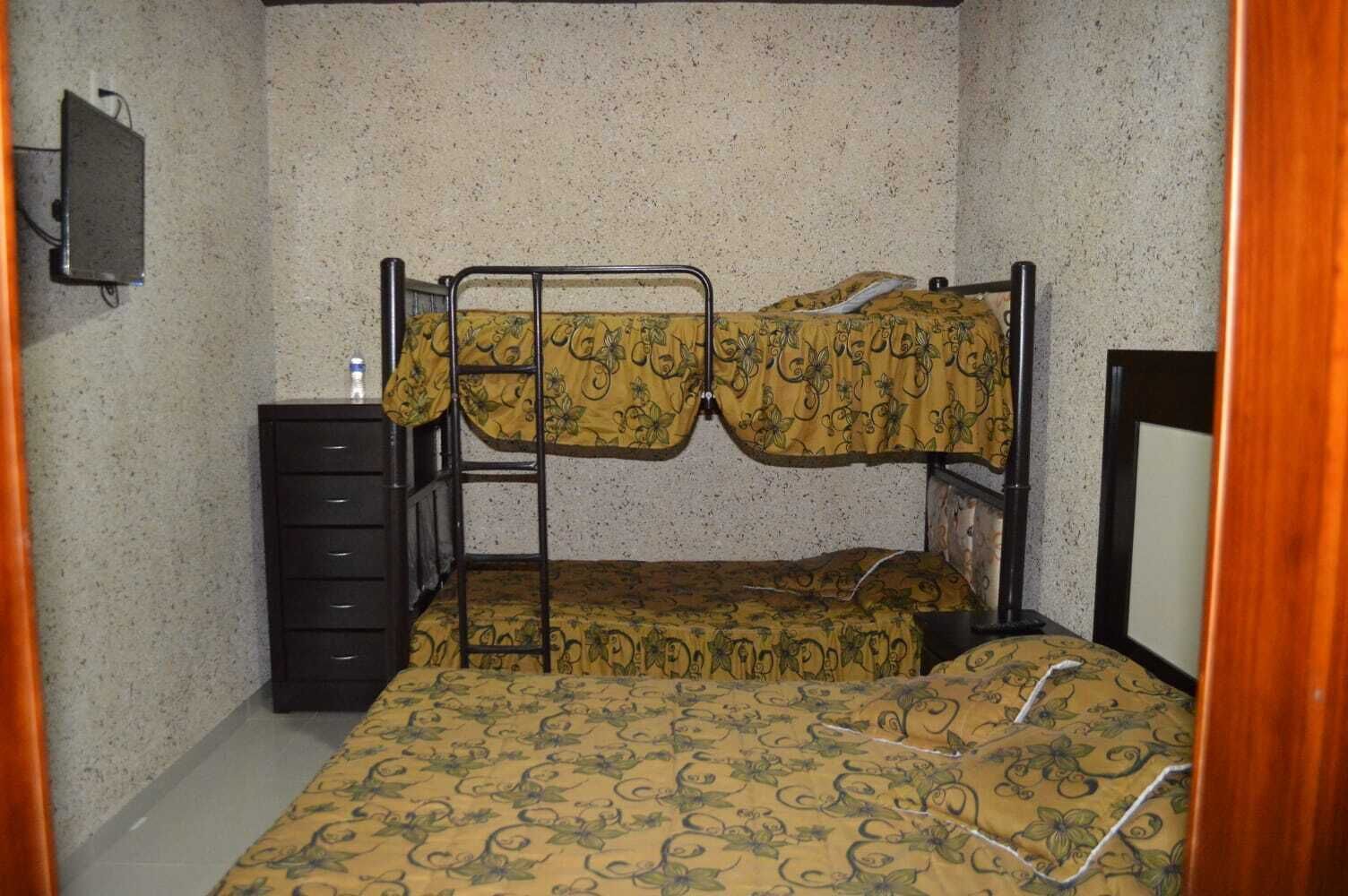 A bunk bed with a patterned bedspread, a dresser, and a television on the wall.