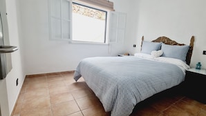 A bedroom with a bed, a nightstand, a window with shutters, and a tiled floor.