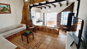 A cozy living room with a sofa, a coffee table with a plant, and a TV. There's a kitchen visible through an open doorway, and a dining area with a table and chairs.
