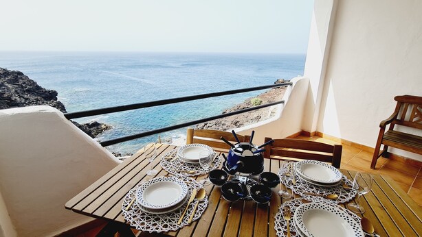 A balcony with a table set for four, overlooking a rocky coastline and the sea.