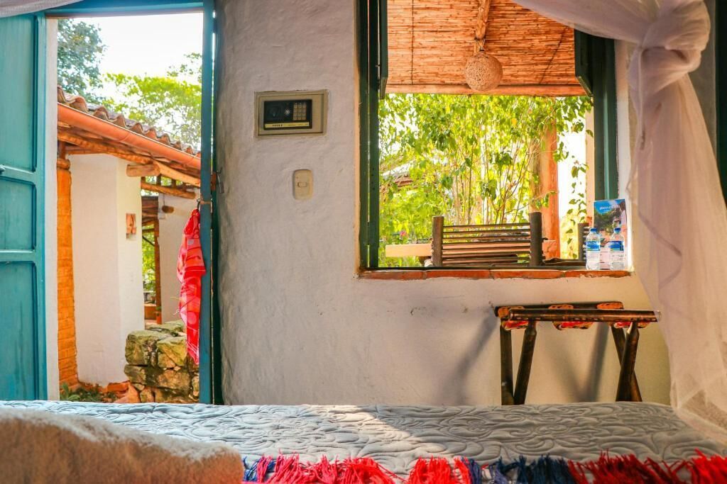 Una habitación con cama, ventana con vista a árboles, una mesita y un banco afuera.