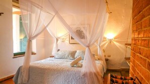 A room with a bed under a canopy, a brick wall, and a small table with a chair. - El Cogollo (Barichara)