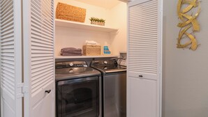 A laundry room with two washers and a dryer, a shelf with towels and a basket, and a wall decoration.