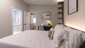 A bedroom with a bed, a desk, and a chair. There is a door leading to another room and a wall with a framed quote.