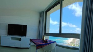 Una habitación con un televisor sobre un mueble blanco, una mesa con mantel a rayas, y vista a una playa y edificios a través de un ventanal grande.