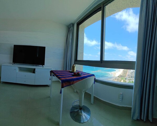 A room with a TV on a white cabinet, a table with a striped tablecloth, and a view of a beach and buildings through a large window.