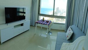 Una sala moderna con televisor de pantalla plana, una mesa de comedor con mantel colorido, un sofá con cojín y un amplio ventanal con vistas a la ciudad y al océano.