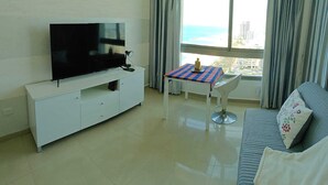 Una sala de estar con un televisor de pantalla plana sobre un mueble blanco, una mesita con mantel a cuadros, una silla blanca y un sofá con un cojín.