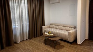 Una sala de estar moderna con un sofá color beige, una mesita redonda con un frutero y un ventanal grande con cortinas marrones.