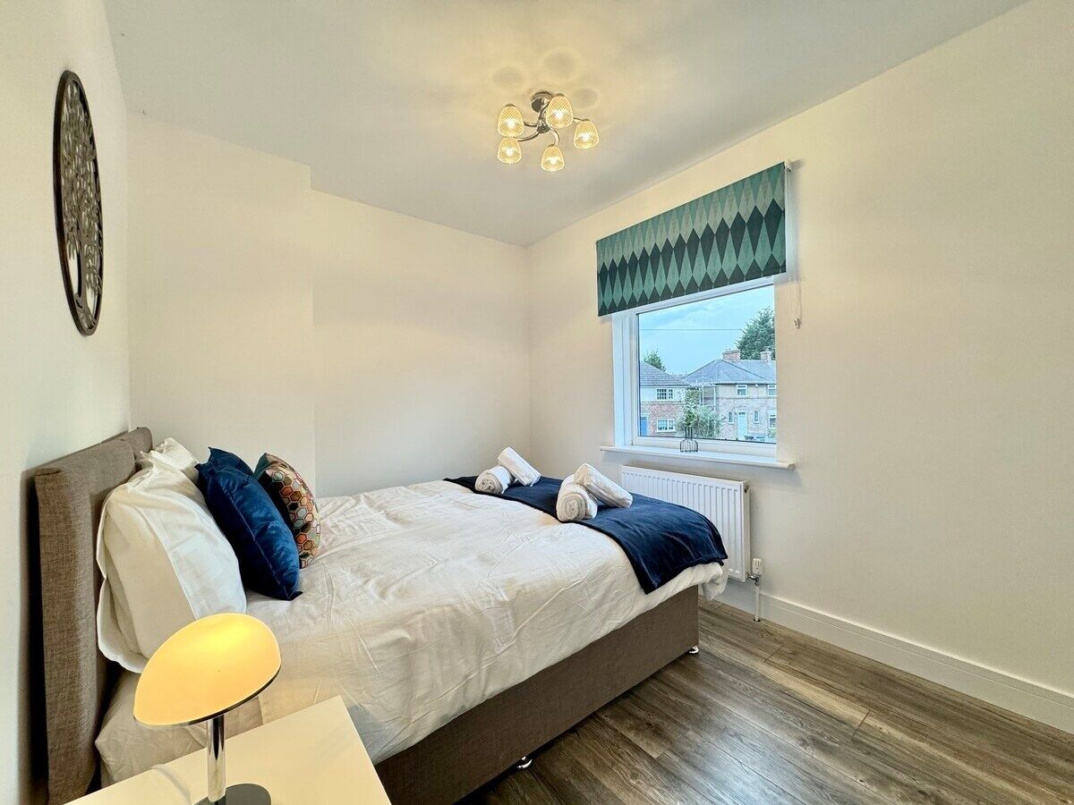 A bedroom with a bed, bedside table, lamp, window, and a view of the outside.