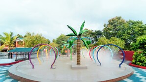 Una fuente de agua con arcos coloridos y una palmera en el centro, rodeada de un suelo a cuadros y follaje.