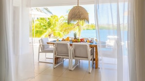 Uma área de jantar com mesa posta para dois, cadeiras brancas, luminária pendente de rattan e vista para uma paisagem tropical através de cortinas leves.