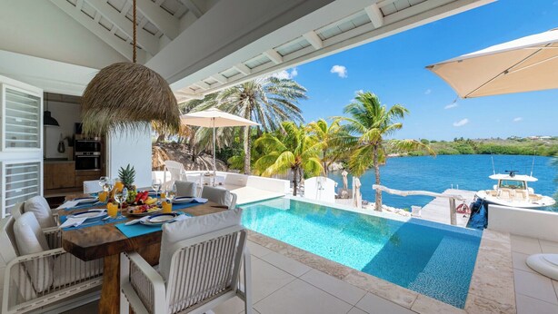 Uma área de refeições à beira da piscina com vista para o mar, um barco ancorado perto e um cenário tropical com palmeiras.