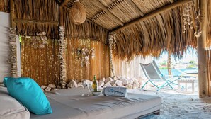 Um quarto à beira-mar com telhado de palha, uma cama com travesseiro, uma garrafa e copos sobre a cama.
