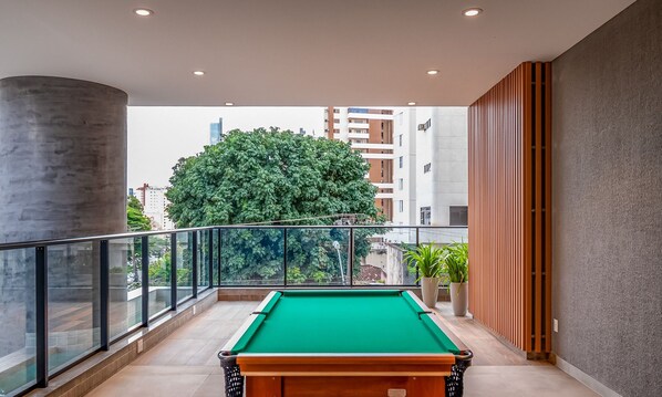 Une table de billard dans un décor intérieur moderne, avec une rambarde en verre, un grand arbre et des bâtiments en arrière-plan.