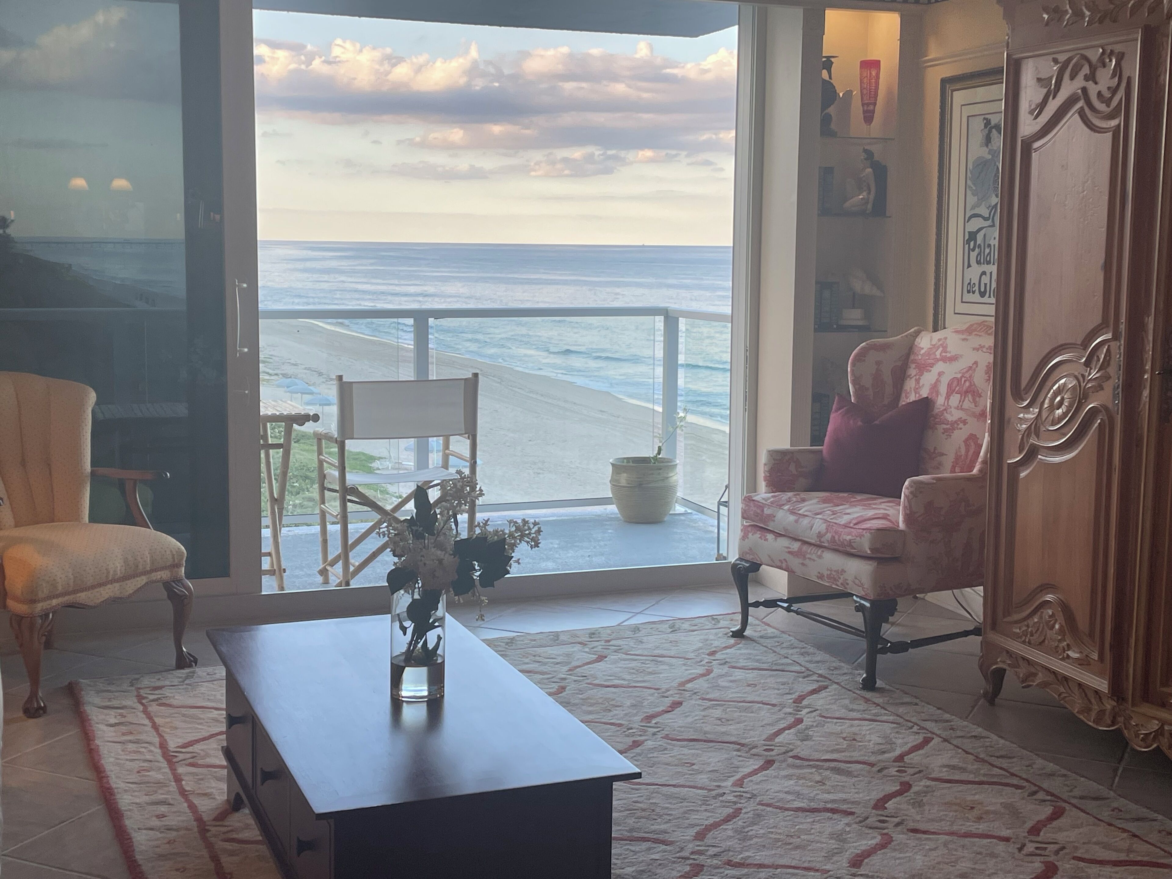 A room with a view of the ocean, a chair, a sofa, a cabinet, and a table with a vase.