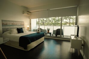A modern hotel room with a large bed, a small table with a chair, a TV, and a balcony with a view of trees. - Sucre Suites (CABA)