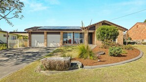 A modern house with a garage, a well-maintained lawn, and a large rock feature in the front yard. - Colloden Beach House by Experience Jervis Bay (Vincentia)