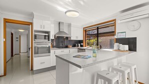 A modern kitchen with white cabinetry, a built-in oven, a microwave, and a large island with bar stools. - Colloden Beach House by Experience Jervis Bay (Vincentia)